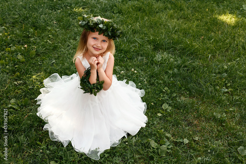 Little blond girl in a wreath of periwinkle on grass background. A child in a white dress sits on the grass