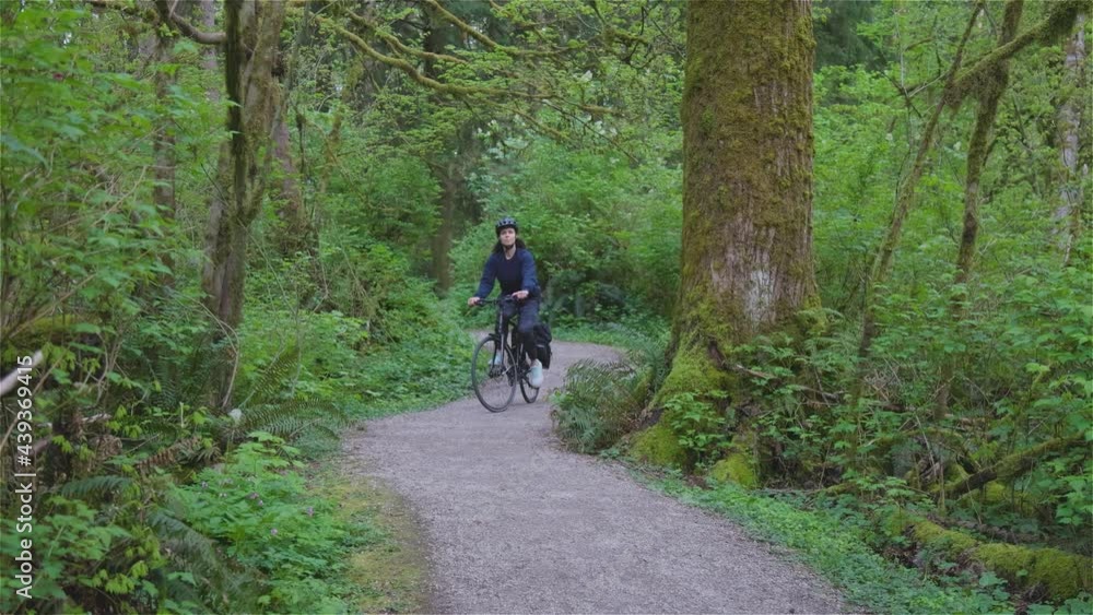 Adult Caucasian Woman Riding a Bicycle on a path in a modern city park. Spring Evening. Taken in Green Timbers Urban Forest, Surrey, Vancouver, British Columbia, Canada. Slow Motion Adult Caucasian Woman Riding a Bicycle on a path in a modern city park. Spring Evening. Taken in Green Timbers Urban Forest, Surrey, Vancouver, British Columbia, Canada. Slow Motion