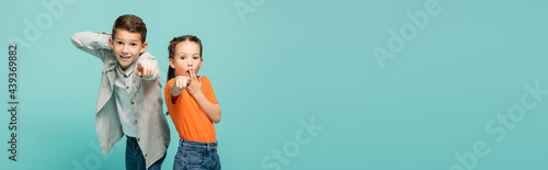 shocked girl in orange t-shirt covering mouth near boy pointing at camera isolated on blue, banner.