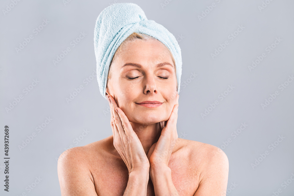 Portrait of smiling senior woman feeling soft skin with hand after spa bath. Mature woman draped in towel looking at camera. Lady in bathrobe after shower isolated over white background.
