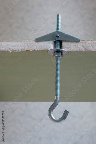 Folding metal dowel hook with spring-loaded wings installed in waterproof sheet of drywall against the background of blurred interior wall, view of section of gypsum fiber in the place of fixation.