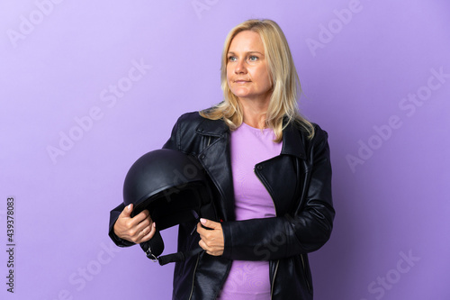 Middle age woman holding a motorcycle helmet isolated on purple background looking to the side