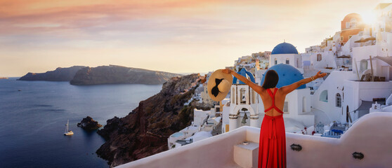 A tourist woman in a red dress enjoys the panoramic view of the village Oia and the caldera on Santorini island, Greece, during summer sunset time