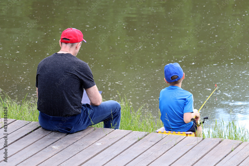 Father and little son on a lake beach. Child sitting with a fishing rod, man with tablet PC, rear view. Summer leisure near the water Father and little son on a lake beach. Child sitting with a fishing rod, man with tablet PC, rear view. Summer leisure near the water