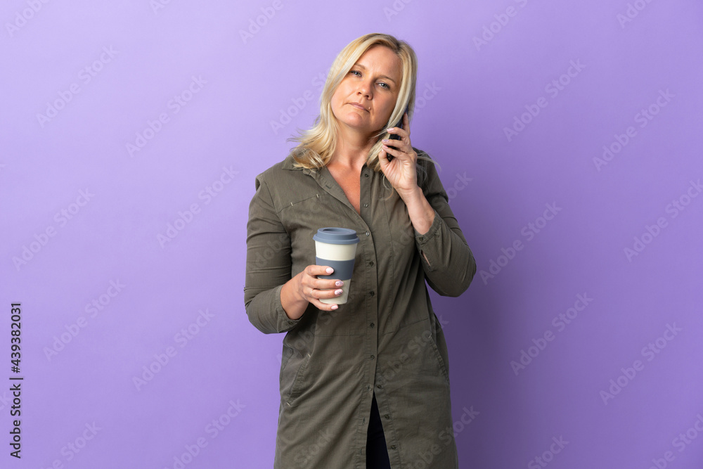 Middle age Lithuanian woman isolated on purple background holding coffee to take away and a mobile Middle age Lithuanian woman isolated on purple background holding coffee to take away and a mobile