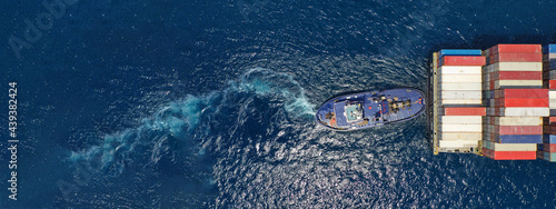 Aerial drone ultra wide top down photo of tow - tug boat assisting by pulling or pushing container ship to anchor in terminal port