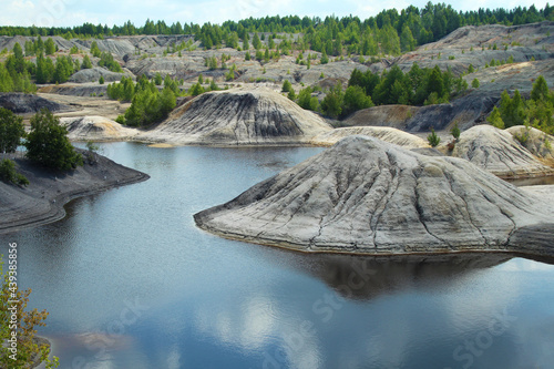 Old quarry for the extraction of clay. A lake among hills of gray and black clay. Trees grow among the hills. In the background there is a green forest. Blue sky and clouds are reflected on the water 