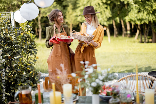 Friends preparing for the festive dinner, two housewifes carrying hot meal and serving the table. A group of young people gathering at backyard of the house in nature