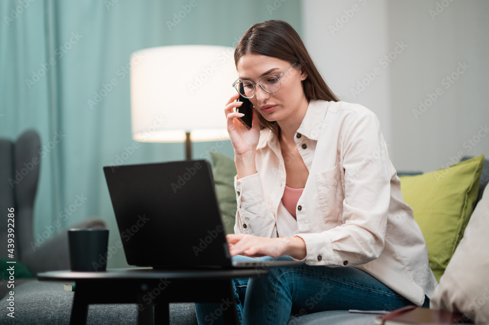 The manager girl holds phone conversations with the client while working remotely from the home office, sitting on the couch in front of a laptop The manager girl holds phone conversations with the client while working remotely from the home office, sitting on the couch in front of a laptop