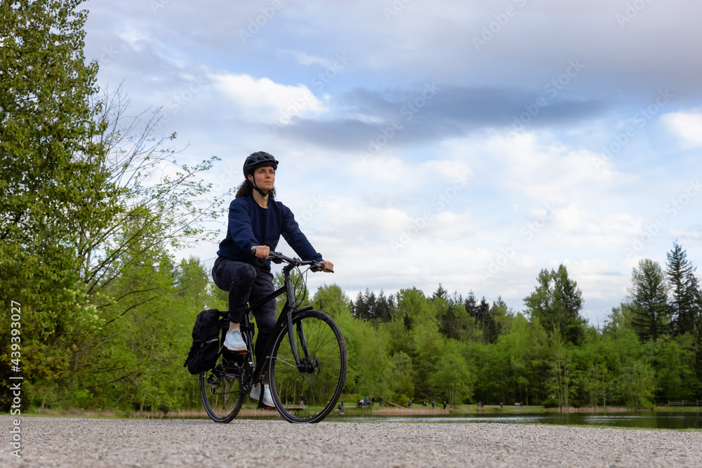 Obraz premium Adult Caucasian Woman Riding a Bicycle on a path by a lake in a modern city park. Spring Evening. Taken in Green Timbers Urban Forest, Surrey, Vancouver, British Columbia, Canada.
