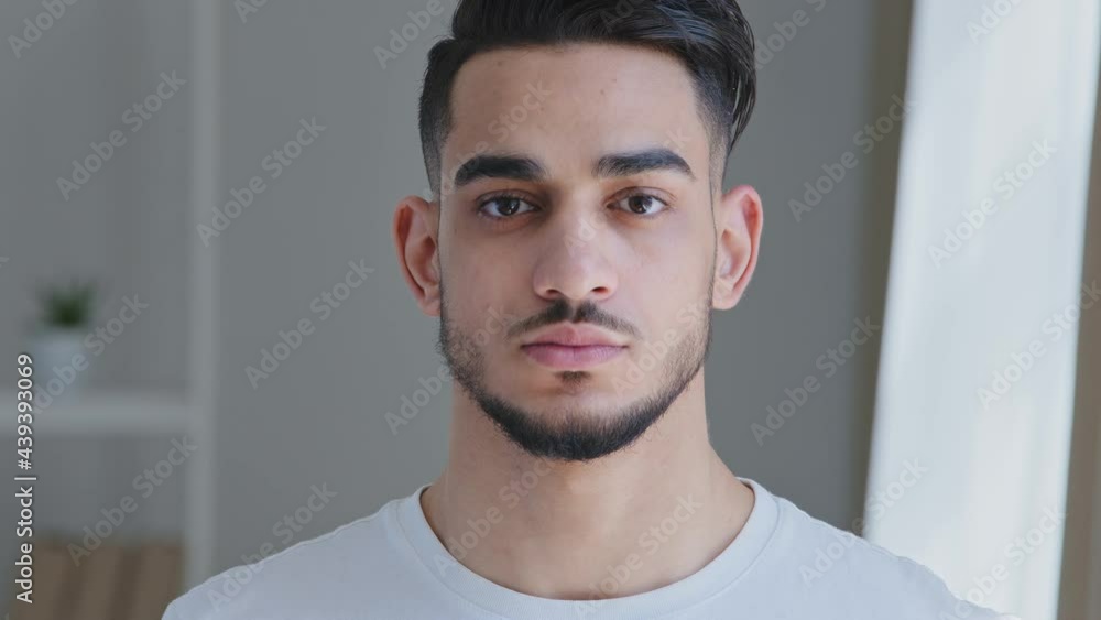 Portrait of serious handsome young adult arabic man with beard in white t-shirt stands posing indoors at home looking at camera. Close-up sad hispanic male ethnic guy brunette freelancer husband model Portrait of serious handsome young adult arabic man with beard in white t-shirt stands posing indoors at home looking at camera. Close-up sad hispanic male ethnic guy brunette freelancer husband model