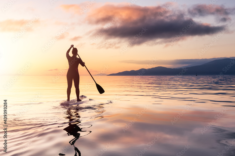 Adventurous woman on a paddle board is paddeling in the Pacific West Coast Ocean. Sunset Sky Art Render. Taken near Spanish Banks, Vancouver, British Columbia, Canada.