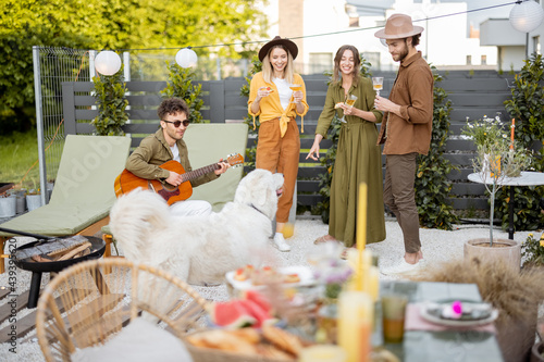 Friends hang out together on a beautiful cottage backyard, playing on a guitar, playing with a dog and eating some snacks. Friends meeting or celebrating outdoors