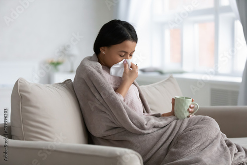 Frustrated sick mixed raced woman wrapped in warm blanket catching cold, suffering from flu illness or infection symptoms, drinking mug of hot herbal tea, blowing nose, taking treatment at home photo