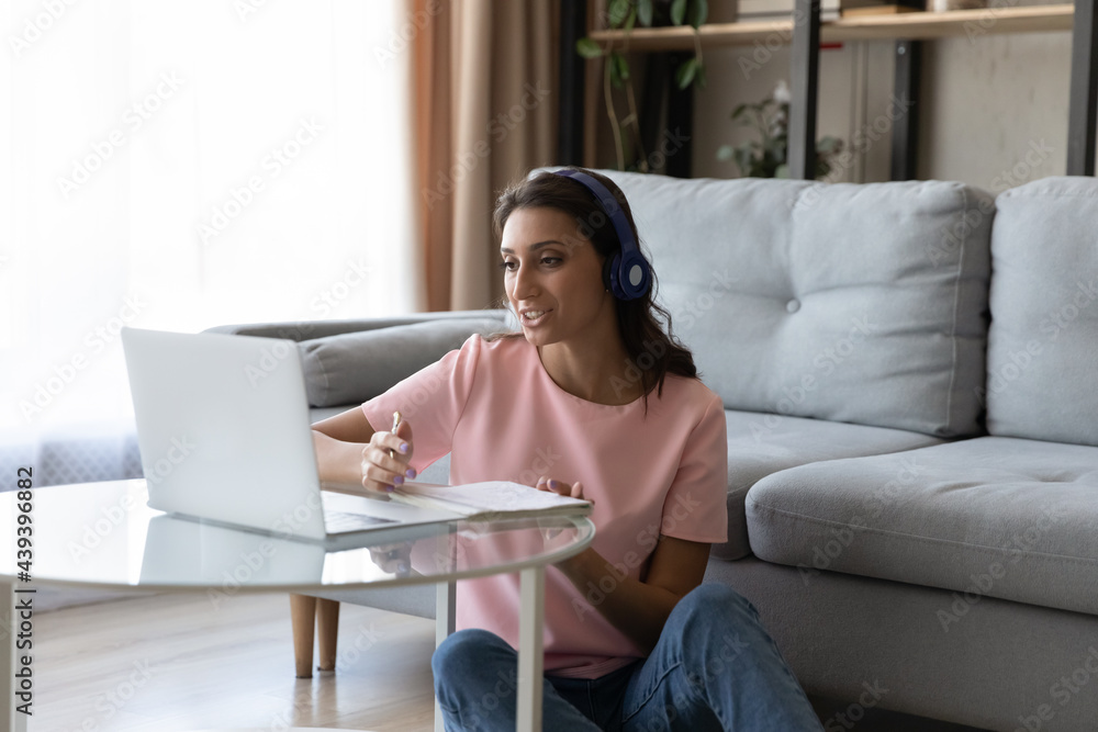 Naklejka premium Smiling Arabian woman in headphones studying online, taking notes, using laptop at home in living room, motivated young female watching webinar, involved in online course, preparing to exam