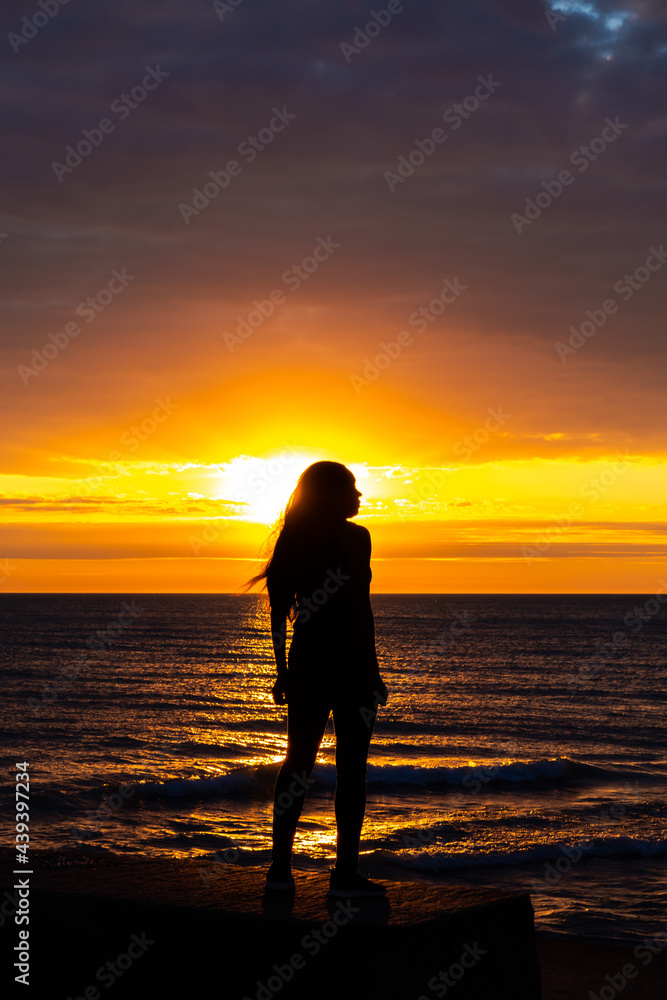 A beautiful African American woman sunrise silhouette on Lake Michigan as she looks out across the water with the orange and yellow clouds reflecting on the water below.