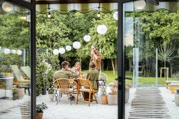 Friends having a festive dinner, gathering together at backyard of the house in nature. View through the window from the inside
