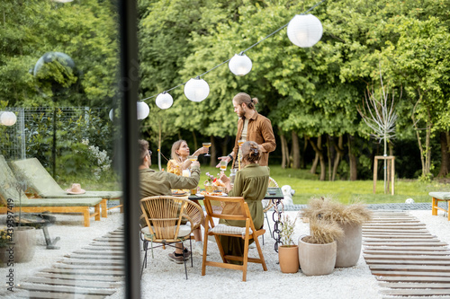 Friends having a festive dinner, gathering together at backyard of the house in nature. View through the window from the inside photo