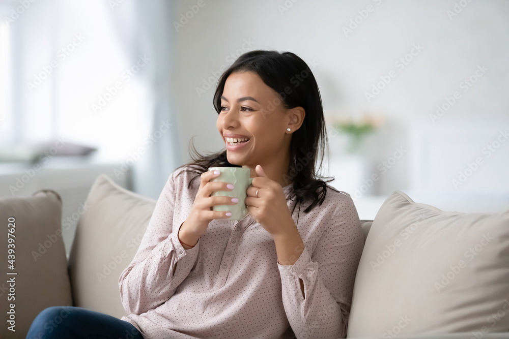Happy black woman enjoying break for leisure at home, relaxing on couch, drinking cup of hot tea or coffee, looking away, smiling, laughing, thinking of good future, watching funny movie on TV Happy black woman enjoying break for leisure at home, relaxing on couch, drinking cup of hot tea or coffee, looking away, smiling, laughing, thinking of good future, watching funny movie on TV