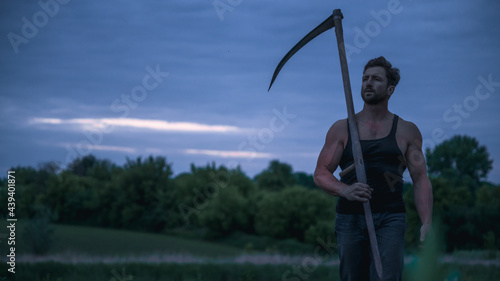 Adult farmer in sleeveless shirt holding scythe in field in morning 