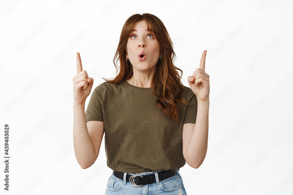 Wow awesome. Impressed girl lift head up, pointing fingers at top advertisement with fascinated face expression, checking out cool promo deal, standing against white background Wow awesome. Impressed girl lift head up, pointing fingers at top advertisement with fascinated face expression, checking out cool promo deal, standing against white background