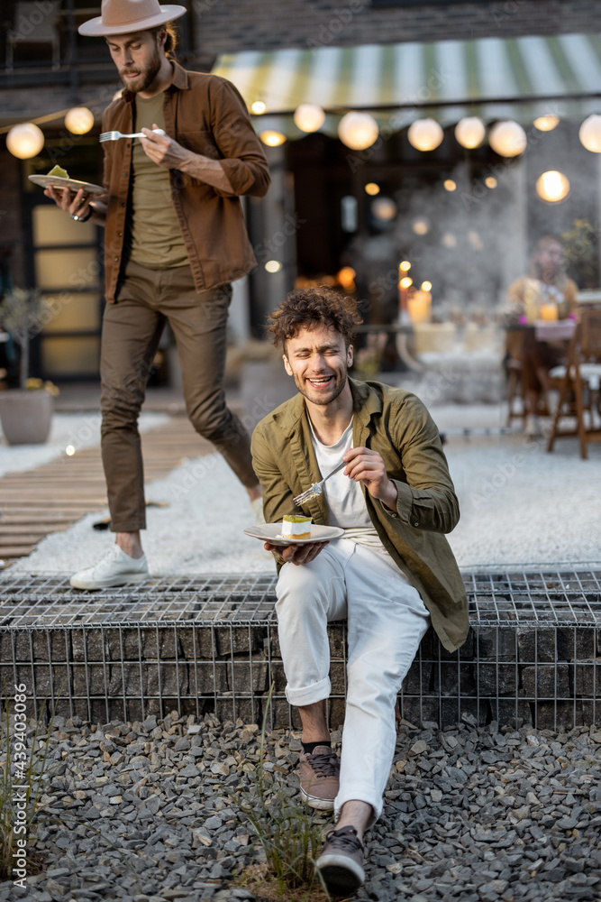 Obraz premium Two male friends eating sweet cakes on a porch of the country house on dusk outdoors. Fun summer time in a country house, male friendship
