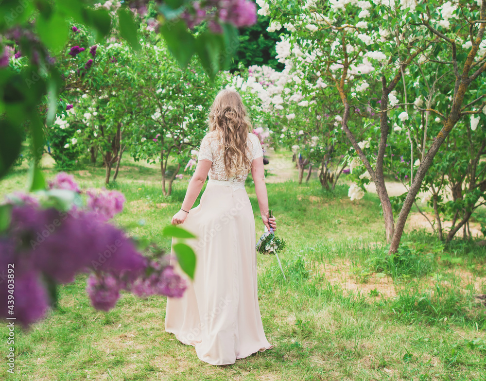 blonde girl in a dress from the back stands with a bouquet around blooming lilacs on the background of a green garden blonde girl in a dress from the back stands with a bouquet around blooming lilacs on the background of a green garden