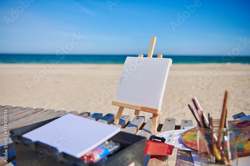 Blank white canvas on a wooden table easel next to paints and brushes on a wooden deck chair on the beach against the background of beautiful blue sea