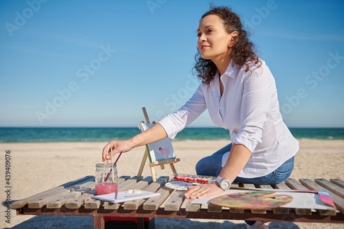 A beautiful young woman sits on a wooden deck chair on the shore of the beautiful sea and writes a picture on canvas.