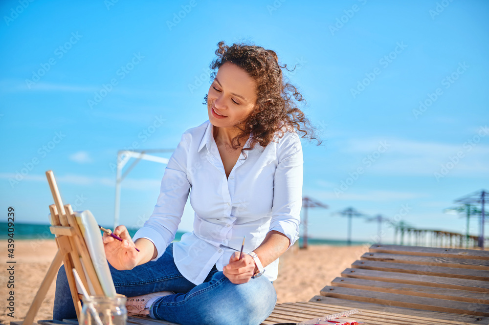 Happy woman artist painting on canvas at the sitting on lonely beach. Art, hobby outdoors .