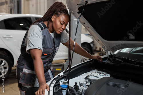 Tired african auto mechanic woman misunderstanding what is wrong with car, looking inside of hood, side view portrait. Young female in auto service. copy space