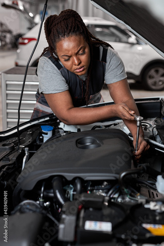Afro American Professional Mechanic Woman Working on Vehicle in Car Service. Female Engine Specialist Fixing Motor, Wearing Overalls and Using a Ratchet. Modern Clean Workshop.