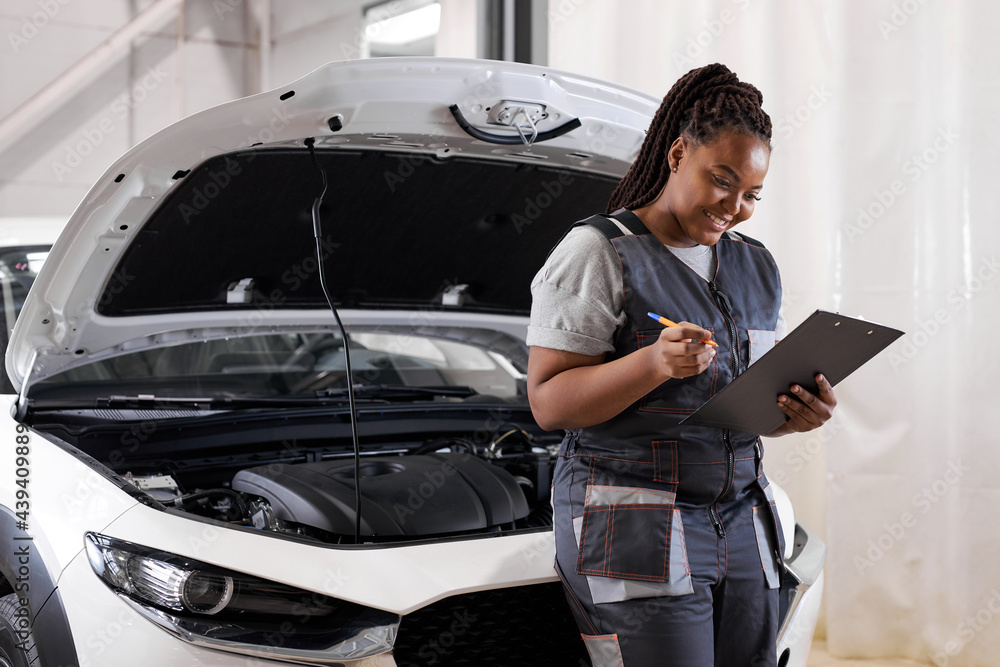 black woman mechanic engineer holding checklist paper and taking notes on clipboard, african woman is standing next to car engine hood, wearing uniform overalls. side view portrait