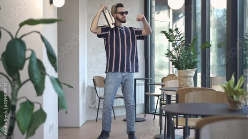 Wide shot of retro dandy man dancing to music from vintage tape recorder taking off sunglasses looking at camera. Portrait of confident Caucasian guy enjoying hobby in coffee bar indoors