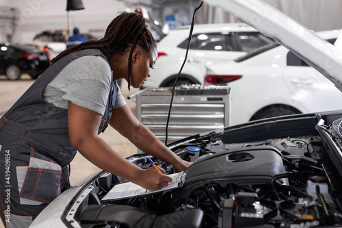 Young afro american woman mechanic holding clipboard checklist at service center repair, side view on confident serious black female at work, thinking. in auto service
