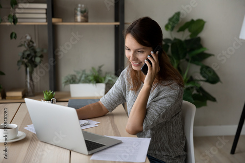 Smiling woman talking on smartphone, looking at laptop screen, confident businesswoman working online, consulting client by phone call or negotiating, discussing project with colleague, multitasking