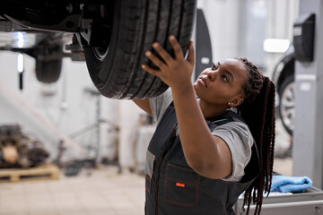 Young afro american mechanic adjusting the tire at repair garage alone, in overalls uniform, black woman working in auto repair shop, focused, raising tire up to lifted car. Side view