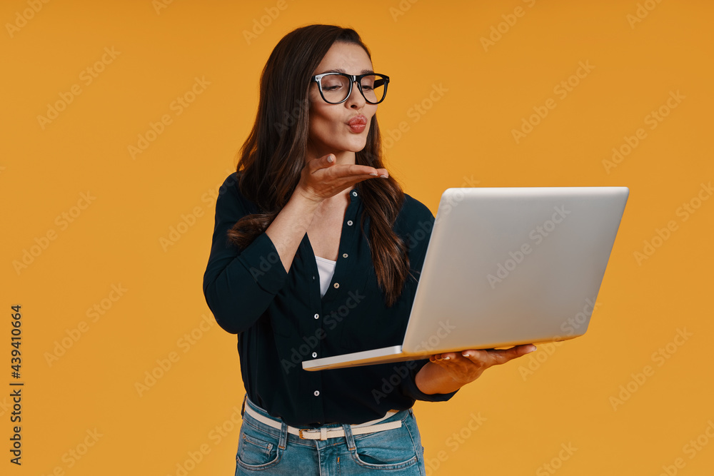 Playful young woman in smart casual clothing using laptop and blowing a kiss Playful young woman in smart casual clothing using laptop and blowing a kiss