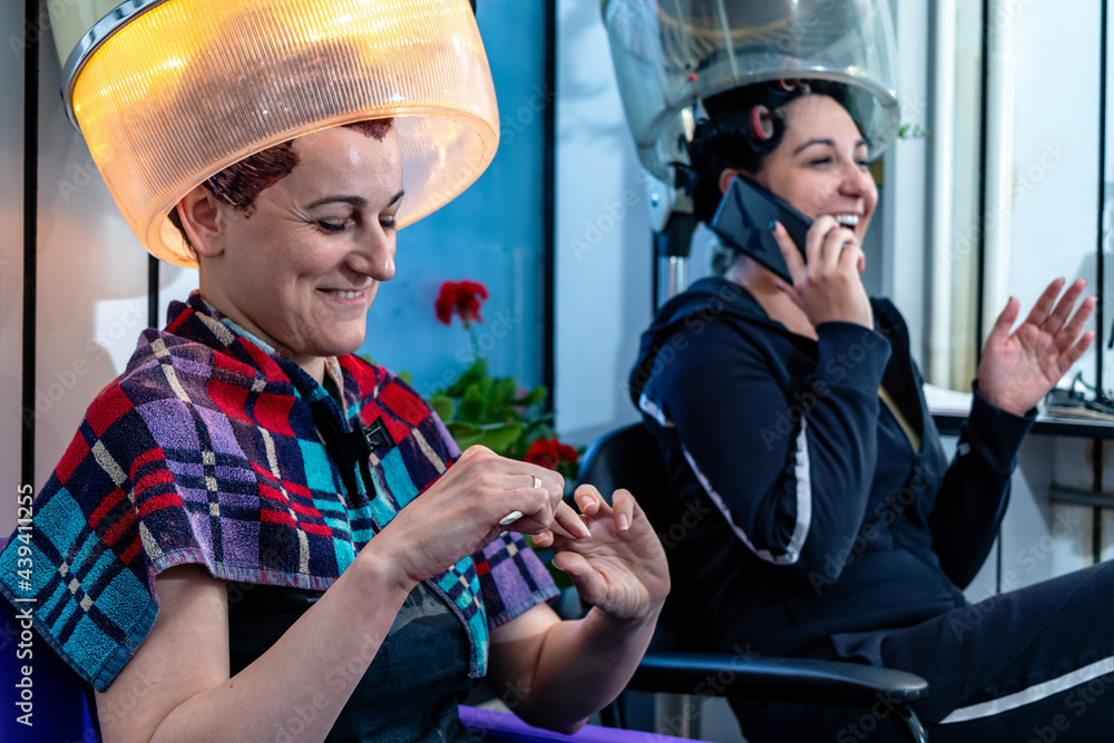 Two female friends at the hairdresser in a beauty salon sitting under a vintage hood hair dryer talking gossiping and laughing while doing their nails. Retro style and fashion. Stock Foto Adobe