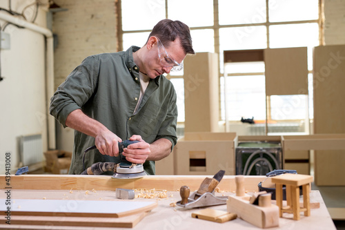 Young male woodworker or joiner working with wood using electric sander in workshop. Joinery work on the production and renovation of wooden furniture. Small Business Concept