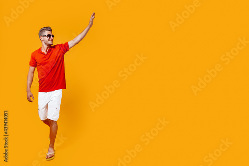 Full length portrait of a handsome man in shorts and red shirt walking, looking sideway raised his hand in greeting. 