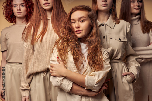 Redhead curly female in casual shirt coat posing at camera standing among female models in the background, portrait. Isolated beige background. fashion, style, trend concept
