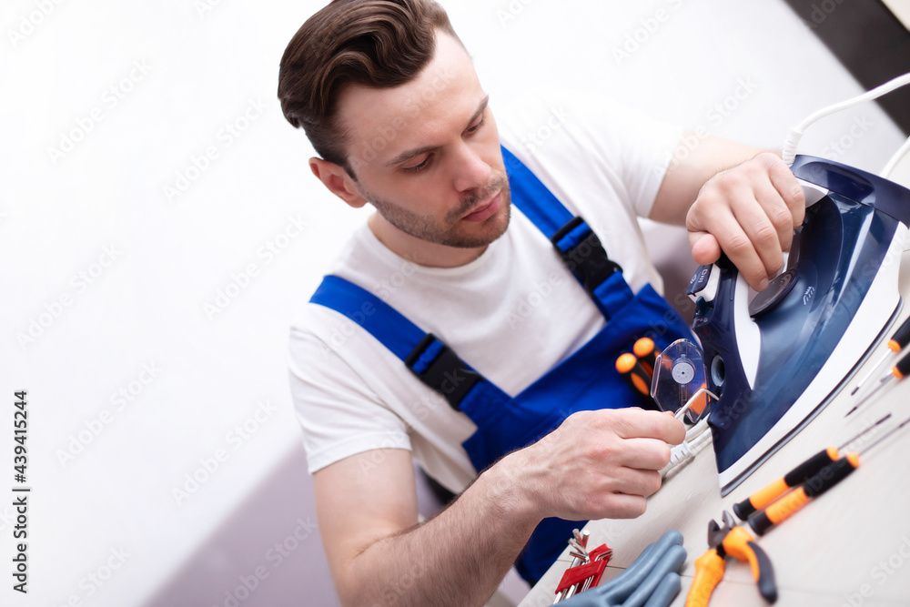 Close up photo of concentrated young man while he is fixing damaged iron on his workplace with tools