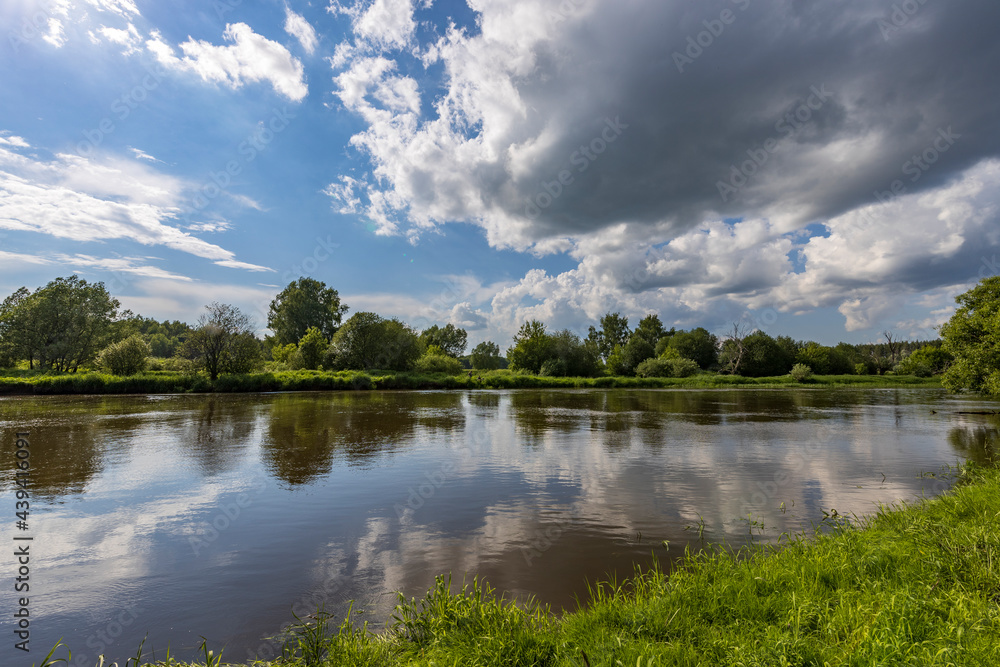 A large cloud is reflected in the river. Rural landscape. Bright green grass on the river bank. Summer landscape with a river and bushes along the river bank.