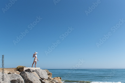 Beautiful blond hair female model clinging to hat while looking out into the large Pacific Ocean from the rocky shoreline.