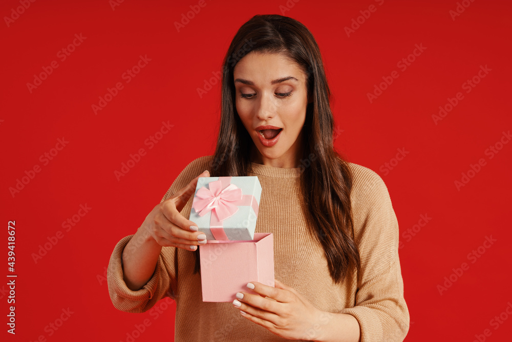 Surprised young woman in casual clothing holding a present while standing against red background Surprised young woman in casual clothing holding a present while standing against red background
