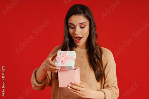 Surprised young woman in casual clothing holding a present while standing against red background © gstockstudio