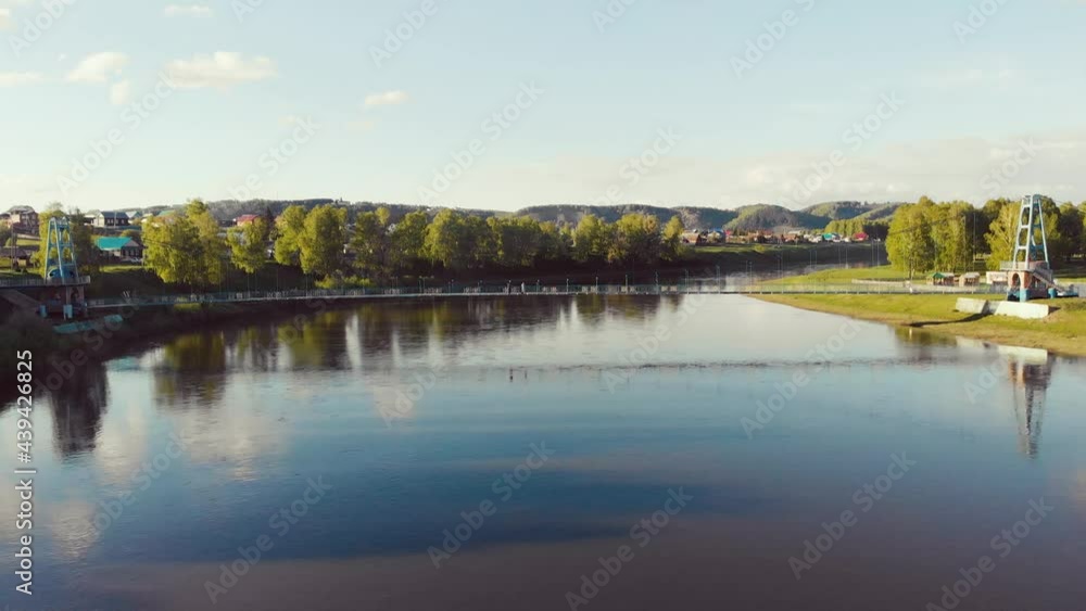 Aerial view of the footbridge over the river, South Ural, Russia. Flying on drone over wide river on the banks of which dense green vegetation. The sun is shining in the blue sky and clouds are moving Aerial view of the footbridge over the river, South Ural, Russia. Flying on drone over wide river on the banks of which dense green vegetation. The sun is shining in the blue sky and clouds are moving