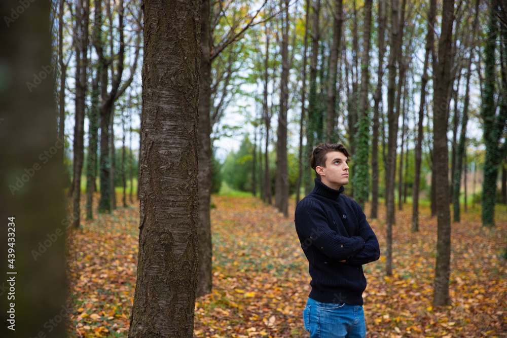 A young man with folded arms looks at the sky and stands in an autumn forest,the leaves are yellow and have fallen from the trees,he wears a wool sweater.Pensive and thoughtful male.Loneliness concept