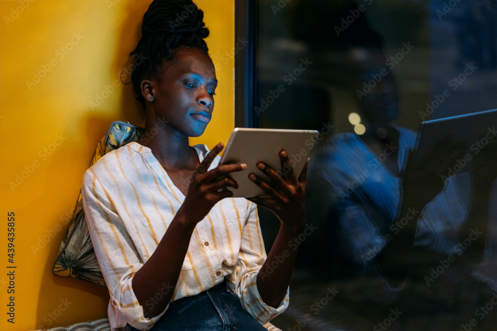 Black woman using tablet at night lying on cushions on windowsill Black woman using tablet at night lying on cushions on windowsill